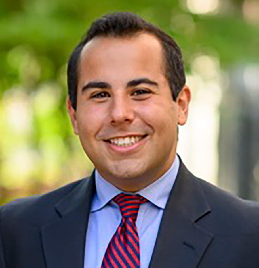 A portrait headshot photograph of Michael Krone (JD candidate and Council of Student Representatives, President) smiling outside in a dark navy blue suit and red plus blue striped patterned colored tie