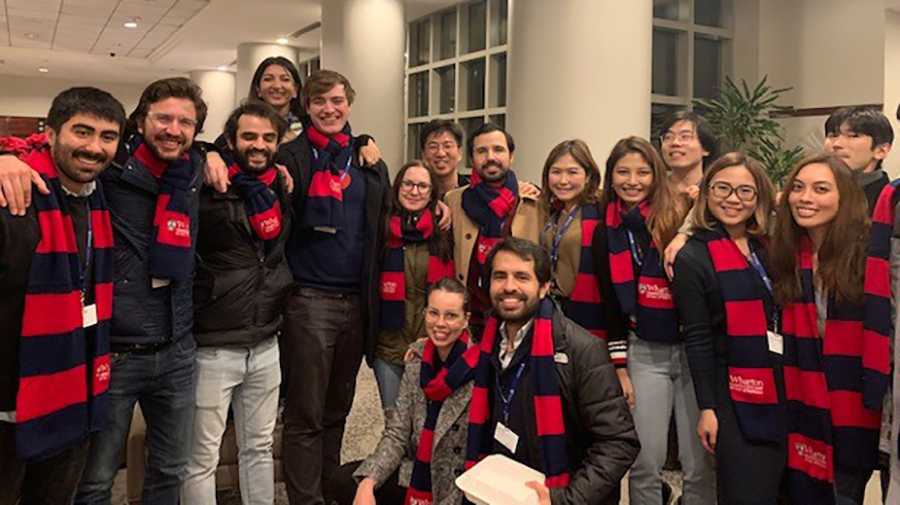 A landscape photograph of Penn Carey Law LLM candidates enrolled in Wharton Business and Law Certificate (WBLC) celebrating the end of their first semester by posing for a picture together all smiling in their dark blue and red scarfs around their necks inside a building